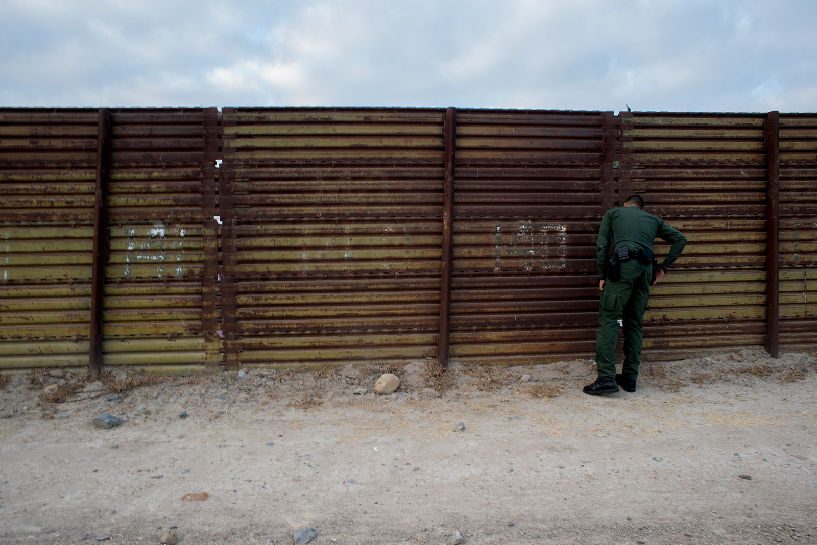 Eduardo Olmos, a Border Patrol public affairs officer, peers through a section of primary fencing between the San Ysidro and Otay Mesa ports of entry in San Diego on Aug. 16, 2017. <em>(Brandon Quester/inewsource)</em>